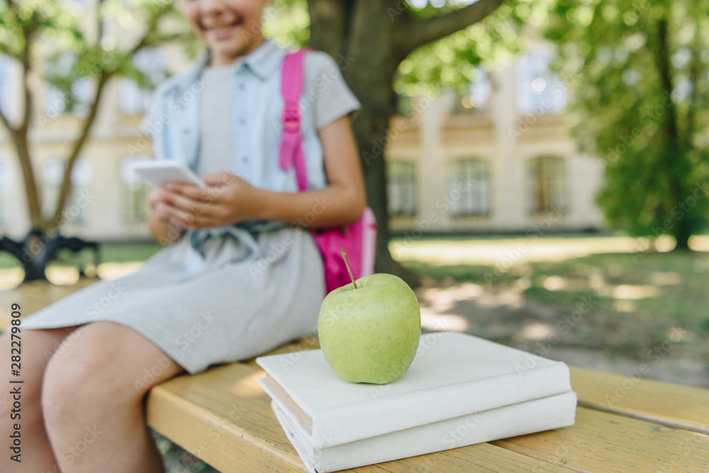 Fototapeta premium cropped view of smiling schoolgirl using smartphone while sitting on bench near books and apple