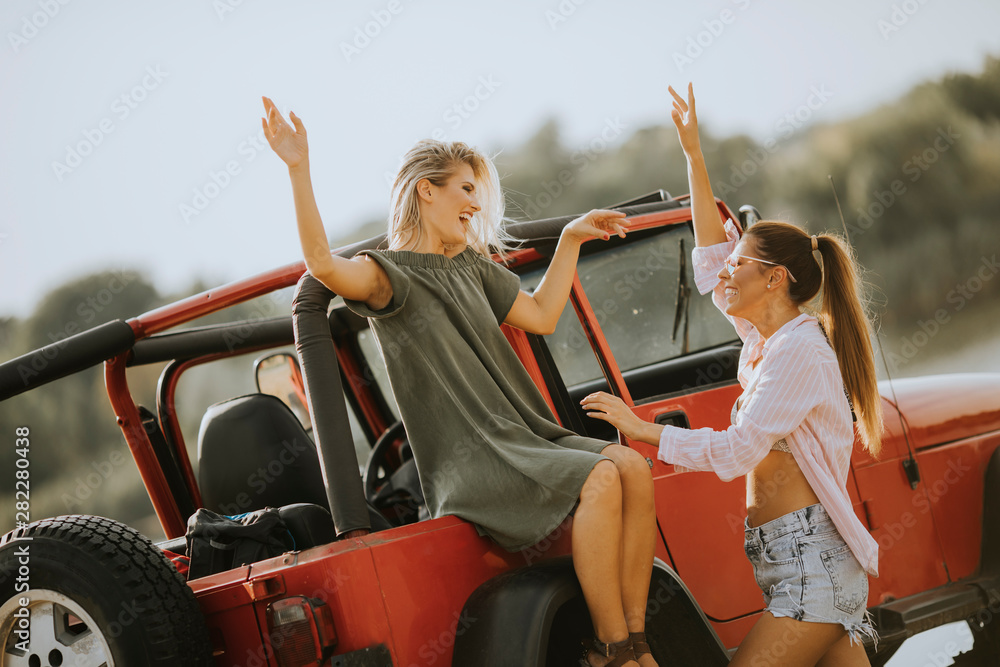 Two young women having fun on hood of a car by the lake Two young women having fun on hood of a car by the lake