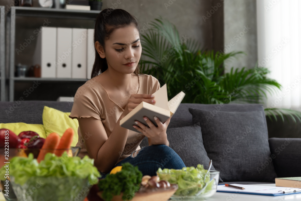 Modern woman love and take care health,sit on sofa,search information,learn to mix vegetable salad in book at home.