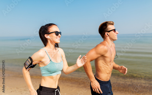 fitness, sport and technology concept - happy couple with earphones and arm bands running along summer beach