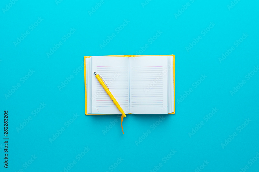© Ruslan Grumble - Top view of opened notebook and yellow pen over it in the centre of turquoise blue background with copy space. Minimalist flat lay image of blank diary and ball-point pen
