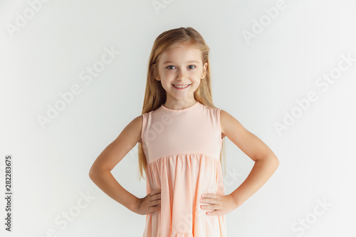 Stylish little smiling girl posing in dress isolated on white studio background. Caucasian blonde female model. Human emotions, facial expression, childhood. Smiling, holding hands on a belt.