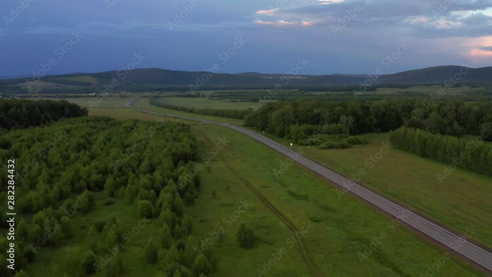 Aerial shot of the area. View of the road on which cars go. In the background is a mountain range. There are clouds in the sky. At sunset, scarlet clouds. Aerial shot of the area. View of the road on which cars go. In the background is a mountain range. There are clouds in the sky. At sunset, scarlet clouds.