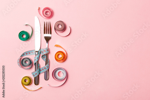 Composition of fork and knife surrounded with colored balled measuring tapes on pink background. Top view of obesity concept with copy space