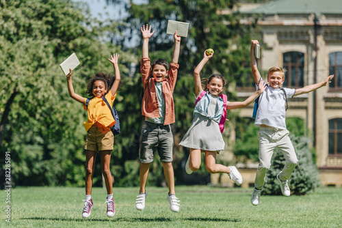 four excited multiethnic schoolkids jumping while holding books on lawn in park