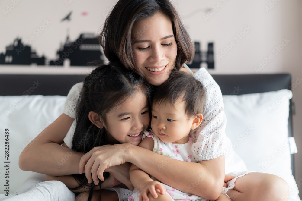 Happy Asian family activity concept. Beautiful mother hugging her children with smile sitting on bed in bedroom. Older sister feeling funny and teasing the baby with happiness. Lovely expression. Happy Asian family activity concept. Beautiful mother hugging her children with smile sitting on bed in bedroom. Older sister feeling funny and teasing the baby with happiness. Lovely expression.