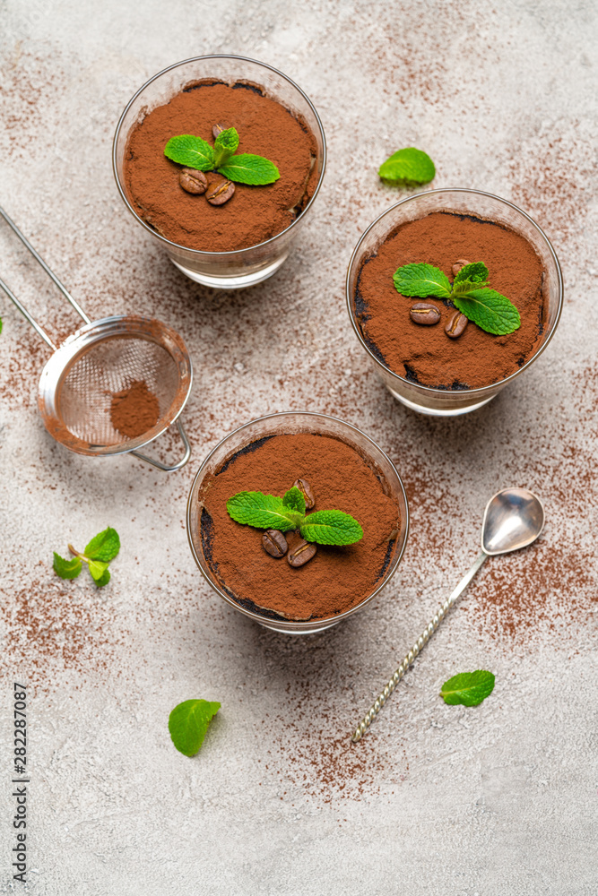 Classic tiramisu dessert in a glass cup and strainer with cocoa powder on concrete background Classic tiramisu dessert in a glass cup and strainer with cocoa powder on concrete background