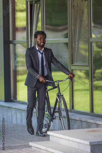 smiling african american businessman walking with bicycle near office building