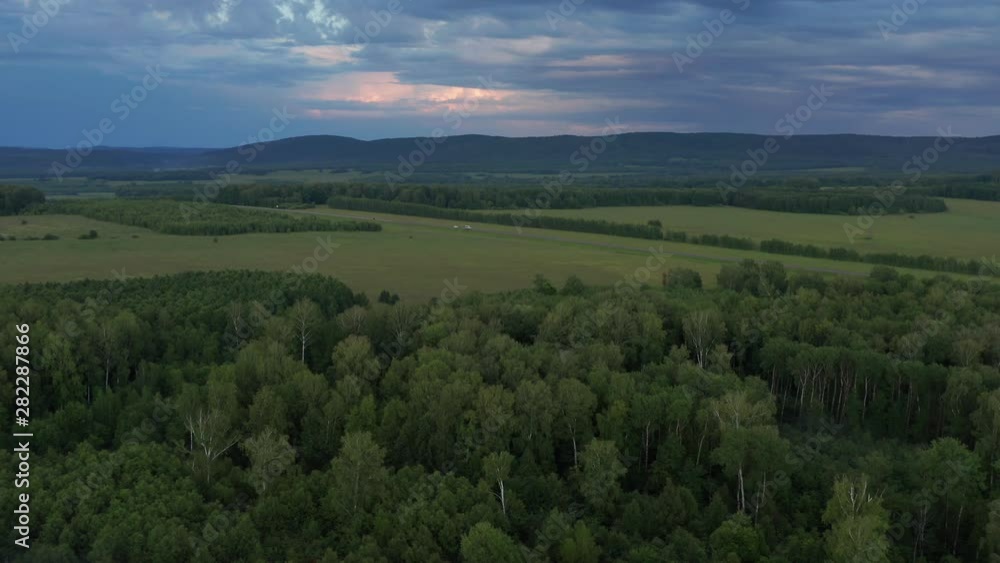 Aerial shooting over dense forest, field and road. In the background are mountains. Evening hours after sunset. Aerial shooting over dense forest, field and road. In the background are mountains. Evening hours after sunset.