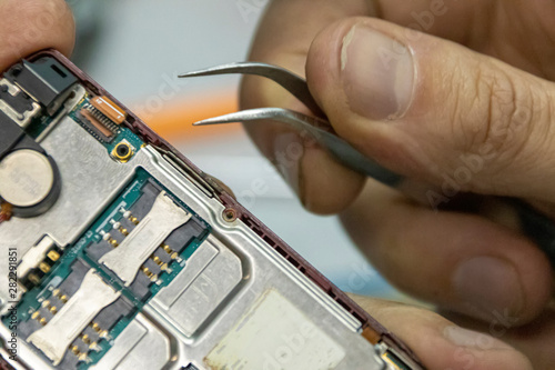 Elderly and young men repair a mobile phone together. They work in a repair shop. They use different tools. They are repair specialists.