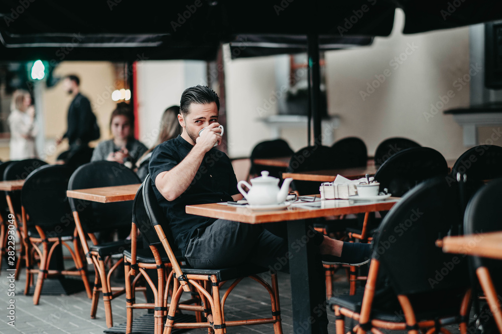 Stylish young brunette man sits in a cafe on the terrace and drinks coffee. Italian street Stylish young brunette man sits in a cafe on the terrace and drinks coffee. Italian street