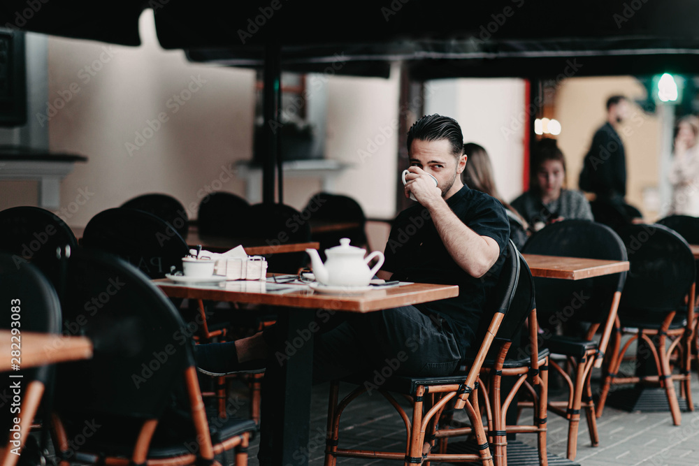 Stylish young brunette man sits in a cafe on the terrace and drinks coffee. Italian street