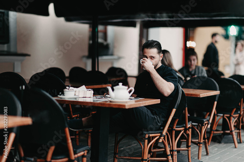 Fototapeta Naklejka Na Ścianę i Meble -  Stylish young brunette man sits in a cafe on the terrace and drinks coffee. Italian street