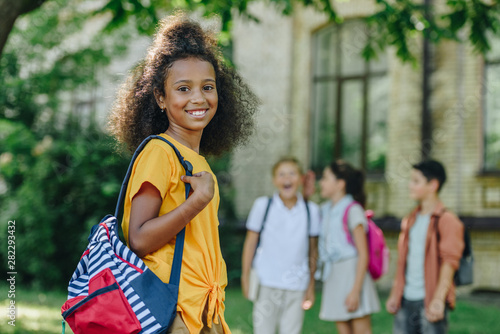 selective focus of cheerful african american schoolgirl with backpack smiling at camera near multiethnic friends
