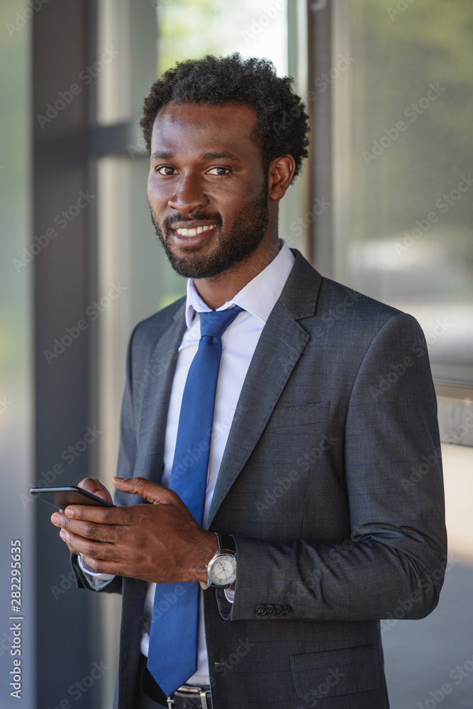 handsome, cheerful african american businessman smiling at camera while using smartphone handsome, cheerful african american businessman smiling at camera while using smartphone