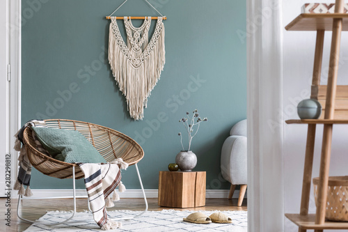 Modern and bohemian composition of interior design at apartment with rattan armchair, wooden cube, plaid, pillows, flowers, macrame, bamboo shelf and elegant accessories. Stylish home decor. Template. photo