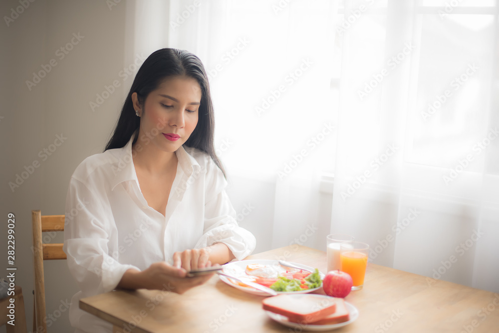 Asian women in white shirt ordering products online with mobile phones in the morning. Happy woman with smartphone and breakfast on the table-Image