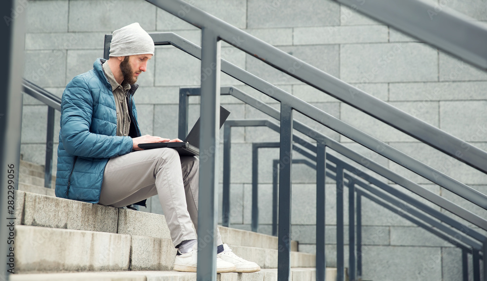 Male freelancer doing remote job using laptop computer. Bearded man working on laptop while sitting  on the steps outdoors at the city.