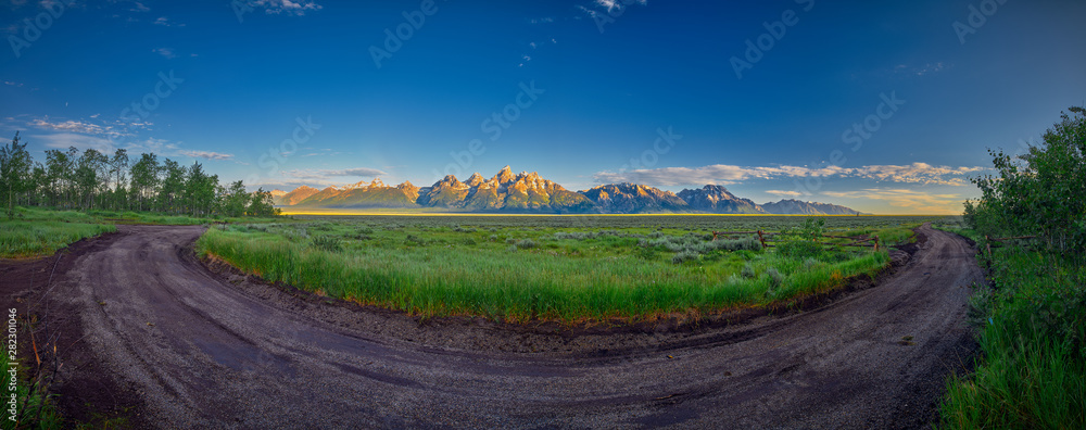 Fototapeta premium Panoramic view of early sun rays on the Grand Teton mountain range with dirt road in the foreground.