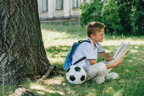 cute smiling schoolboy reading book while sitting on lawn near soccer ball