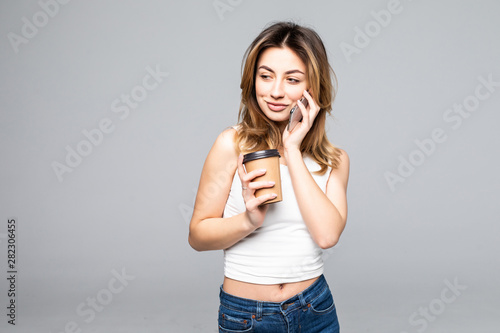 Young woman posing isolated over grey wall background talking by mobile phone drinking coffee.