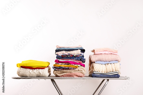 Stack of clean freshly laundered, neatly folded women's clothes on pressboard. Pile of shirts, dresses and sweaters on striped ironing board, concrete wall background. Copy space, close up, top view.