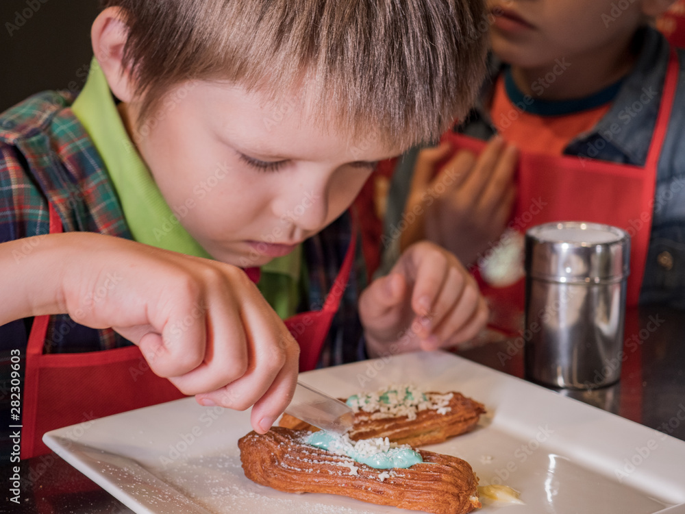 Funny children in red aprons eat eclairs cooked at master class. Confectionery education. Cookies, cakes, sweet cream, puff pastry. Vanilla, mint and confectionery powders Funny children in red aprons eat eclairs cooked at master class. Confectionery education. Cookies, cakes, sweet cream, puff pastry. Vanilla, mint and confectionery powders