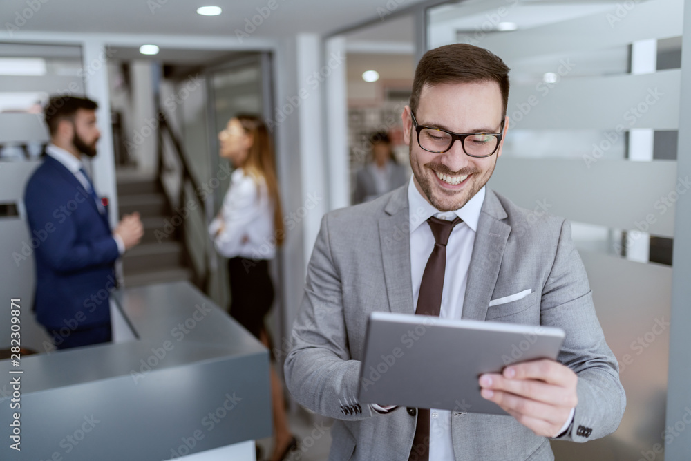 Portrait of handsome Caucasian smiling unshaven businessman in suit and with eyeglasses standing in hall and using tablet. In background his colleagues chatting. Portrait of handsome Caucasian smiling unshaven businessman in suit and with eyeglasses standing in hall and using tablet. In background his colleagues chatting.