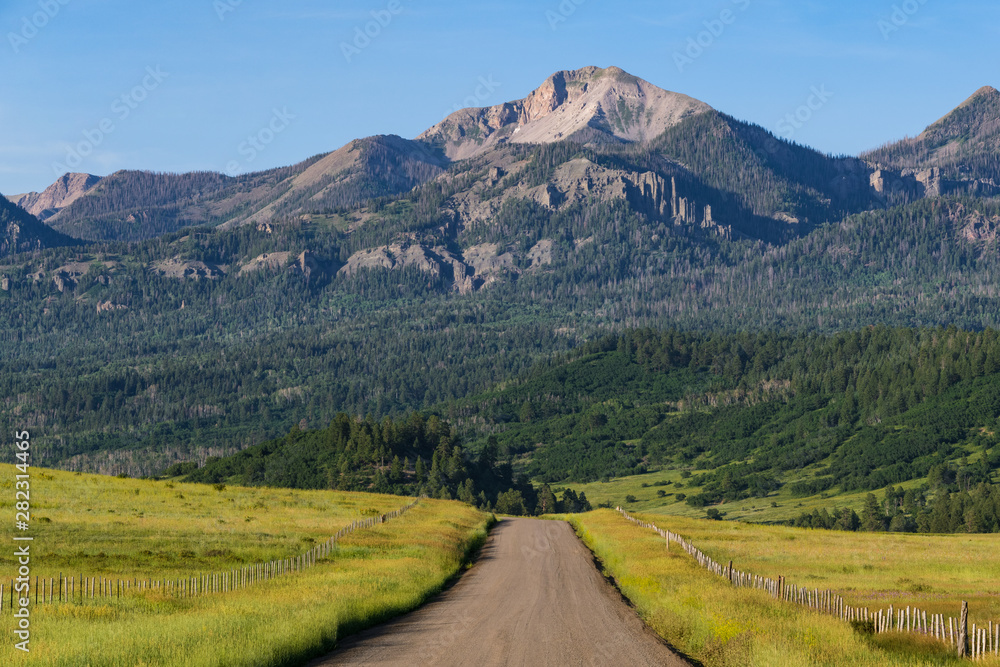 Rural scene of a road and fences through grassy meadows and fields below a high mountain peak - the Rocky Mountains near Pagosa Springs, Colorado