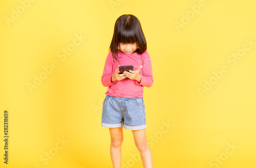 Curious little Asian girl playing games on her mobile phone isolated over yellow background.