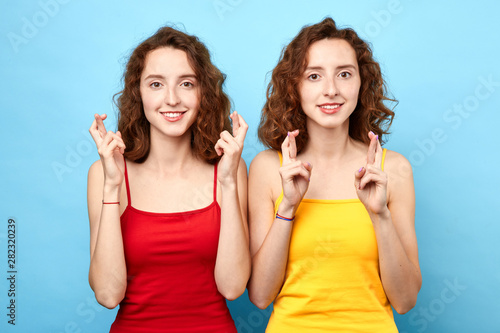 Fototapeta happy charming brunette women wears tops, smiles broadly, keeps fingers crossed, hopes for good luck, isolated against blue background with copy space for your text or advertisment