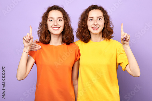 Happy beautiful twins girls point up isolated on blue background, two sisters showing something above their heads , advertisement, place for text, body language