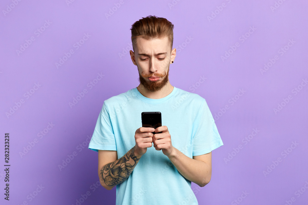 Upset stressed man with funny beard and moustache holding cellphone, received sms, message with bad news, isolated on blue background. Human face expression. emotion, feeling. close up portrait Upset stressed man with funny beard and moustache holding cellphone, received sms, message with bad news, isolated on blue background. Human face expression. emotion, feeling. close up portrait