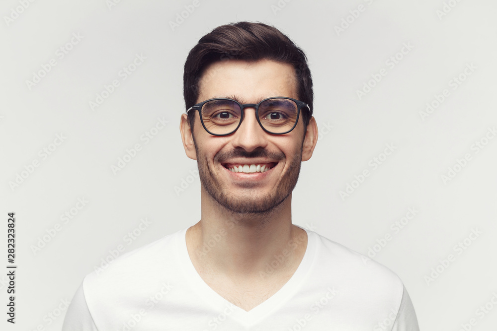 Eyewear fashion. Portrait of smiling handsome young man in casual white t-shirt and trendy eyeglasses, looking at camera, isolated on gray background Eyewear fashion. Portrait of smiling handsome young man in casual white t-shirt and trendy eyeglasses, looking at camera, isolated on gray background
