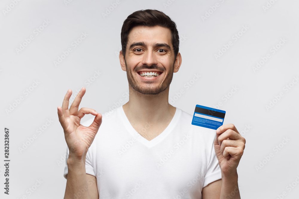Portrait of young smiling man in white t-shirt, holding credit card and showing okay sign, isolated on gray background Portrait of young smiling man in white t-shirt, holding credit card and showing okay sign, isolated on gray background