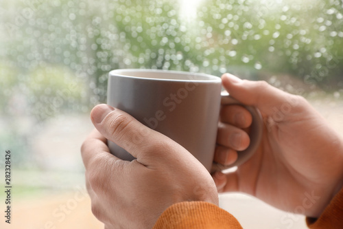 Young man with cup of coffee near window indoors on rainy day, closeup