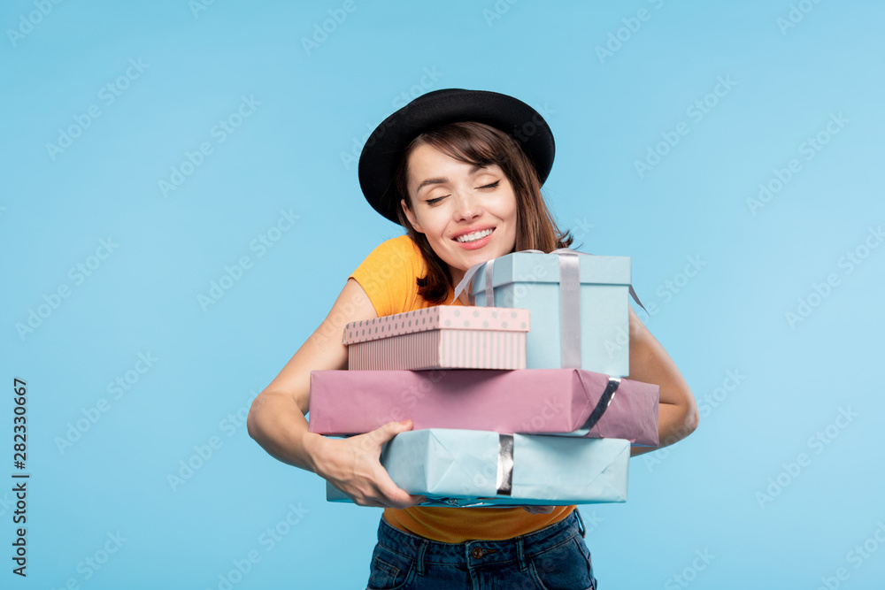 Young cheerful woman in hat embracing stack of wrapped gifts
