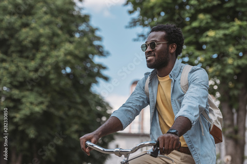 handsome african american man in sunglasses looking away and smiling while riding bicycle