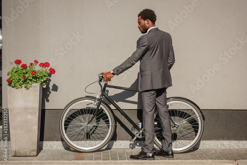 Fototapeta Naklejka Na Ścianę i Meble -  african american businessman in suit standing with bicycle near wall and red flower in flowerpot