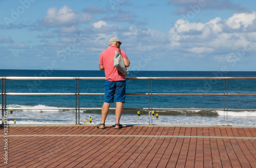 Fototapeta Naklejka Na Ścianę i Meble -  Senior Man in Golden and Glorious Aged Looks From Top Seafront at Activity of Surfer Beginners on the Ocean Coast. View of Beautiful Horizon Line Between Clear Sea Water & Blue Sky in Sunny Day. Spain