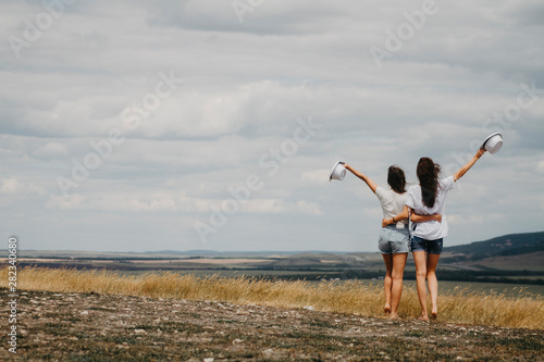Two girls hug each other at the waist and raise their hands up against the backdrop of a landscape of mountains.