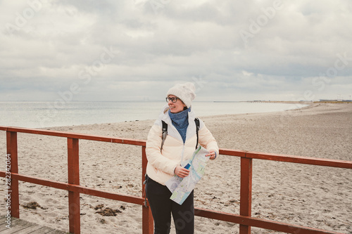 Canvas Print Caucasian woman in hat and jacket with backpack in winter sits on wooden pier on beach near North Sea