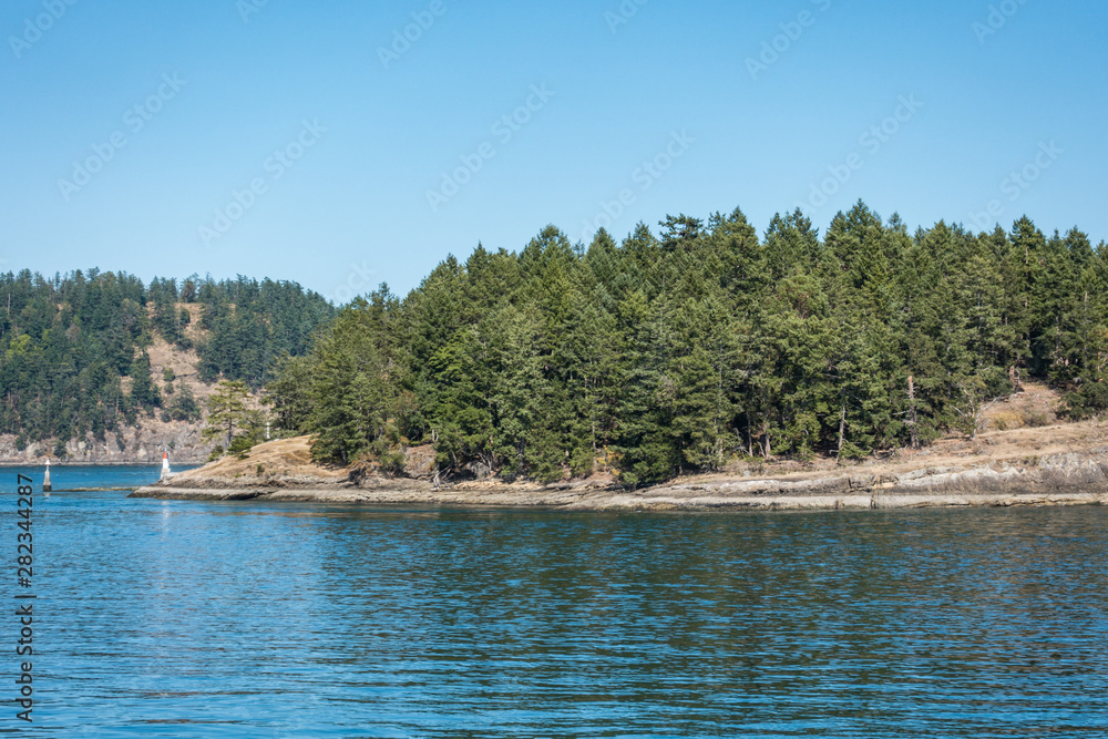 Fototapeta premium view of coast line covered with dense green forest on the island over the horizon under blue sky on a sunny day on the ocean and a small light house on the edge of the island.
