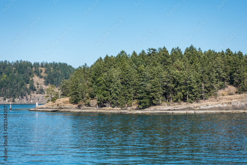 view of coast line covered with dense green forest on the island over the horizon under blue sky on a sunny day on the ocean and a small light house on the edge of the island.