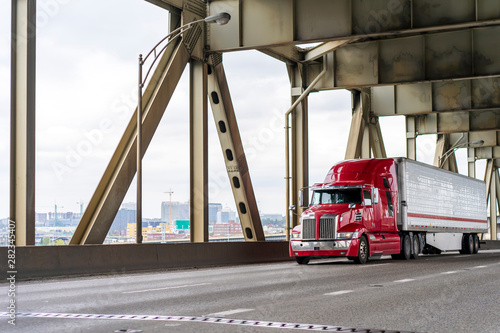 Bright red big rig semi truck transporting cargo in refrigerator semi trailer moving on the two levels bridge across Willamette River in Portland