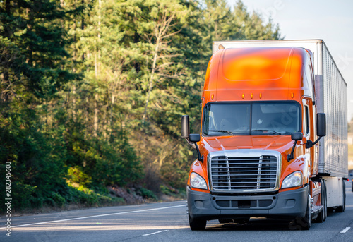 Orange big rig semi truck transporting cargo in semi trailer running on the road with trees