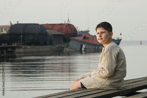 A boy in a linen shirt sits on the edge of a wooden pier amid flooded houseboats © Sergey