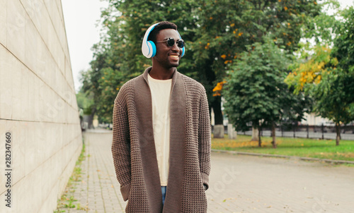 Cool modern african man in wireless headphones listening to music wearing brown knitted cardigan walking in autumn city park in rain photo