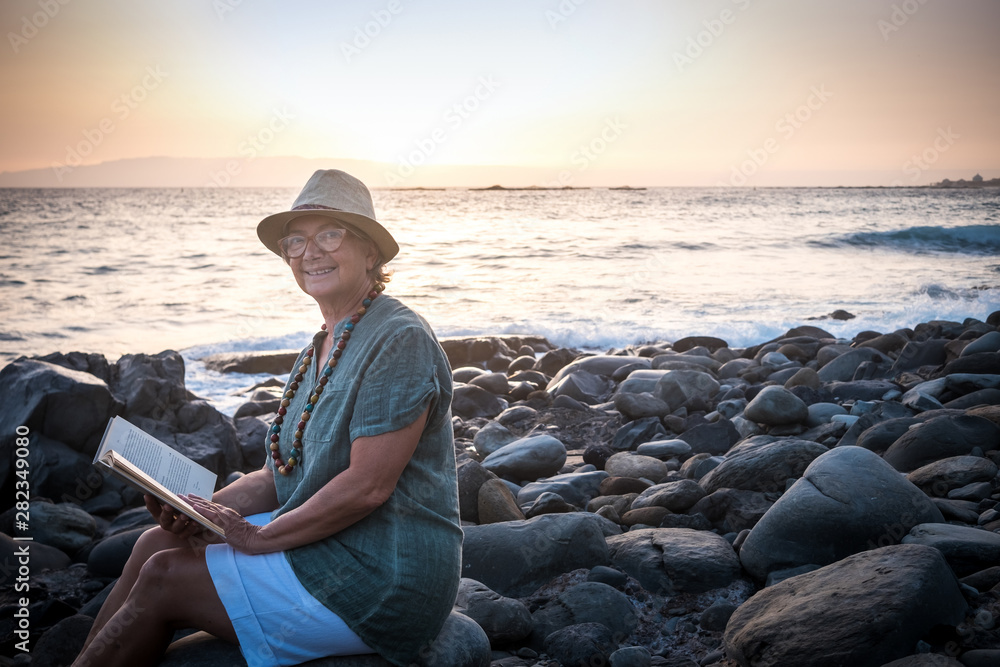 Smiling senior woman with hat sitting on the pebble beach reading a book. Positive moment of relax for the retired grandparent. Background of ocean and sky with the profile of an island