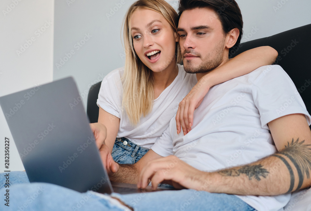 Young thoughtful brunette man and pretty emotional blond woman in white T-shirts using laptop together in bed at home Young thoughtful brunette man and pretty emotional blond woman in white T-shirts using laptop together in bed at home